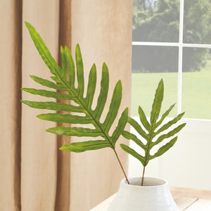 A HARE'S FOOT STEM 34" by NAPA HOME AND GARDEN, featuring elongated fern leaves, is elegantly placed in a white vase on a light wooden table near the window. In the background, a curtain frames the tranquil view of greenery outside.