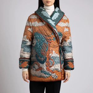 A person wears the SABINA SAVAGE - THE SNOW LION QUILTED JACKET from SABINA SAVAGE ENGLAND, featuring a colorful, intricately designed nature-themed pattern with trees, sky, and animals. Reminiscent of a reversible silk twill jacket, it features a wide collar, contrasting orange inner lining, and large pockets. The person is shown from the shoulders down.