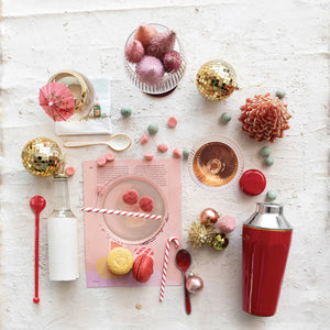 Flat lay of festive cocktail ingredients on a white background, highlighting the 22 oz. enameled stainless steel cocktail shaker by Creative Coop, a gold-rimmed glass with a pink drink, disco balls, colorful sweets, macarons, red spoon, and striped straw. A small cactus and napkin with a drink umbrella add flair.