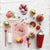 Flat lay of festive cocktail ingredients on a white background, highlighting the 22 oz. enameled stainless steel cocktail shaker by Creative Coop, a gold-rimmed glass with a pink drink, disco balls, colorful sweets, macarons, red spoon, and striped straw. A small cactus and napkin with a drink umbrella add flair.