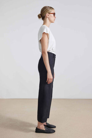 In a minimalist setting, an individual confidently poses against a neutral background, wearing sleek sunglasses, a white T-shirt, the APIECE APART - CLASSIC MERIDA PANT in black high-waisted wide-legged style, and stylish black loafers.