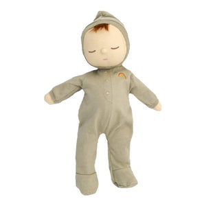 An adorable DOZY DINKUM DOLL by OLLI ELLA USA, featuring soft brown fabric skin, closed embroidered eyes, and short black hair, is dressed in a charming yellow onesie adorned with a rainbow design on the chest. Perfect as a first toy, the doll is lying on a white, quilted surface.