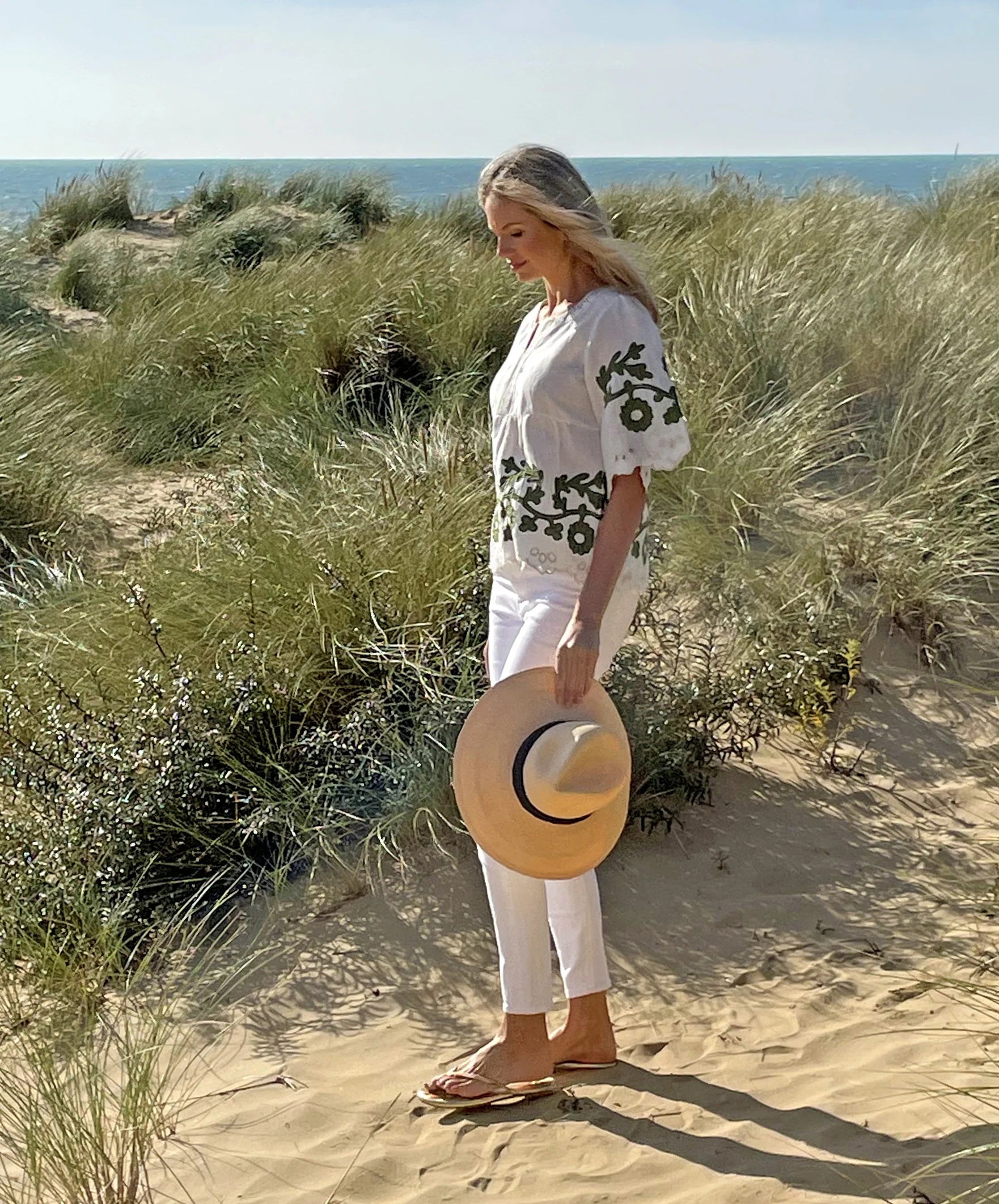 A woman with long blonde hair stands on a sandy beach with tall grass and the ocean in the background. She is wearing the ROSE & ROSE - ALASSIO TOP, a flattering empire line, white linen blend top adorned with daisy-inspired appliqué, paired with white pants and holding a straw hat.