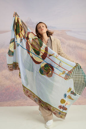 A woman with long blonde hair, in a light blue shirt and dark pants, smiles in front of a scenic backdrop, draping the elegant, hand-embroidered STORIATIPIC - ASTER WOOL SCARF around her shoulders.