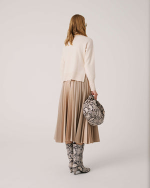 A person with long hair wearing sunglasses poses against a plain background. They wear a cream-colored sweater, a SFIZIO pleated mix media skirt in beige faux leather, and matching knee-high snakeskin boots. They hold a snakeskin-patterned handbag.