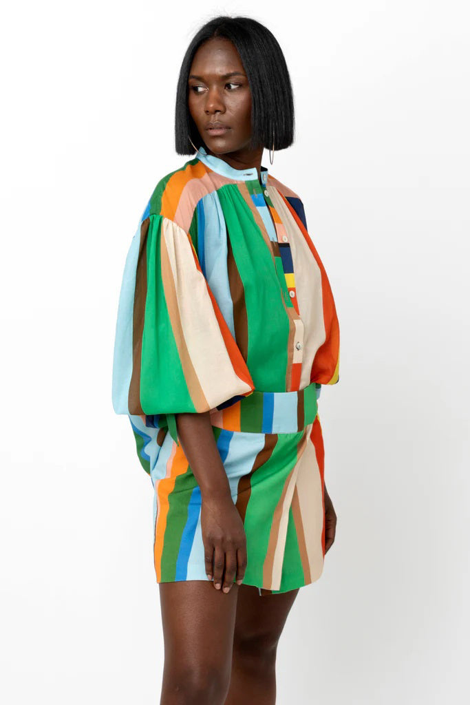 A person stands against a white background, wearing the HOPE FOR FLOWERS Wrist Tie Shirt in a blue and white geometric pattern, paired with a short, multicolored striped skirt. They have shoulder-length hair and are wearing sunglasses. Their hands rest by their sides.