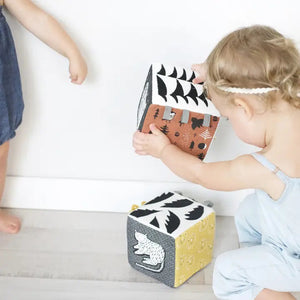 Introducing the WEE GALLERY - WOODLAND SOFT BLOCK by WEE GALLERY, a soft fabric block adorned with a whimsical design perfect for stimulating a baby's developing senses. One side features a black and white squirrel holding a nut, while other sides showcase trees, stars, and owl illustrations in black, white, and brown. Small grey fabric tags protrude from the top for added sensory exploration.