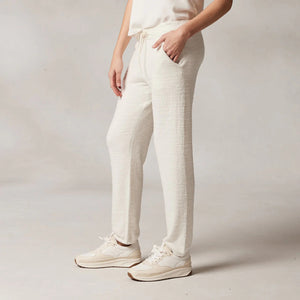 A person stands against a plain background, wearing a white sleeveless top and KASHWERE's RELAXED JOGGER in an ivory color. The joggers are made from lightweight, breathable Kash-ease yarn and have pockets where they’ve placed their hands. The individual completes the look with white sneakers.