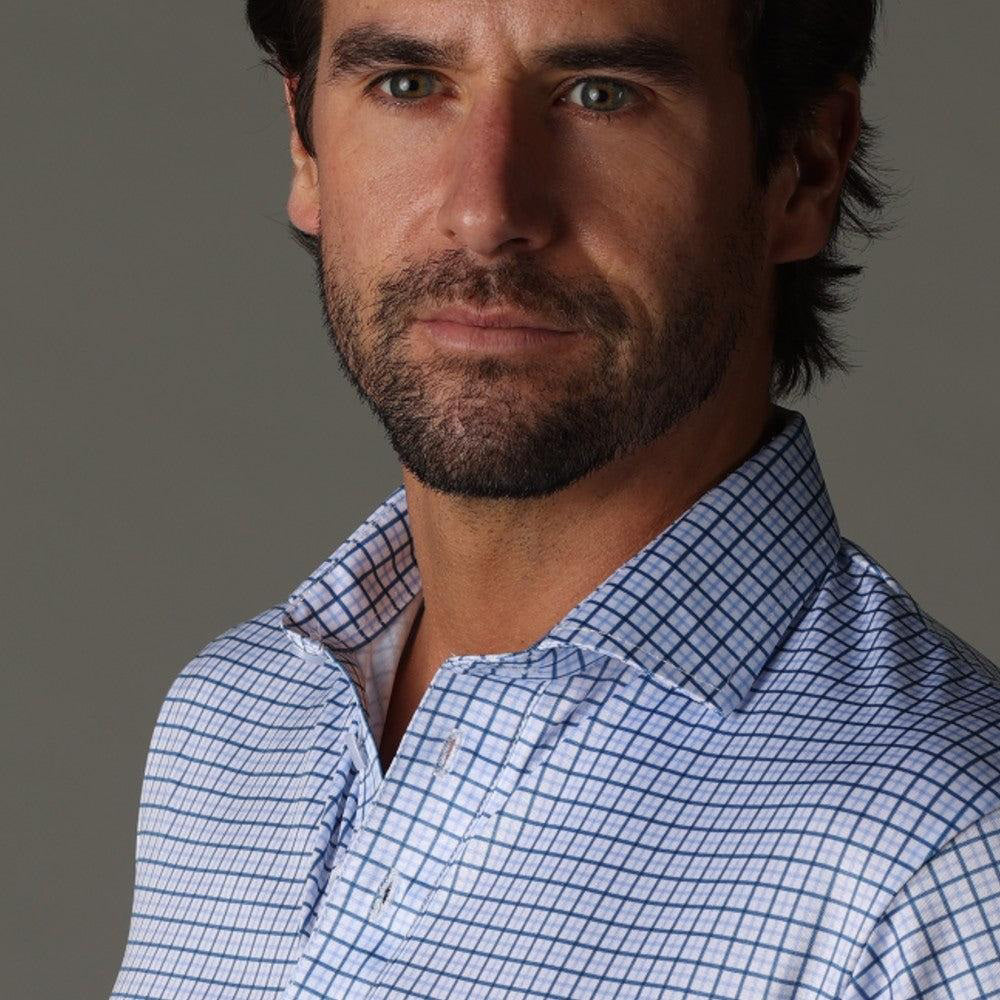 A man with dark hair and a beard wears the COLLARS AND CO - SEMI SPREAD COLLAR POLO IN BLUE TATTERSAL under a light brown sweater, standing against a gray background and looking to his right with a serious expression.