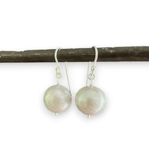 A pair of ANN LIGHTFOOT Freshwater Pearl Drop Earrings, showcasing round, lustrous grey freshwater pearls suspended from delicate sterling silver earwires, is pictured against a simple, dark background.