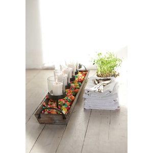 The CREATIVE COOP VOTIVE HOLDER serves as a decorative centerpiece, consisting of a rectangular wooden tray with metal handles. It includes five glass votives, each elevated on black metal stands and surrounded by small pebbles at the base—ideal for adding fireplace ambience to any room.