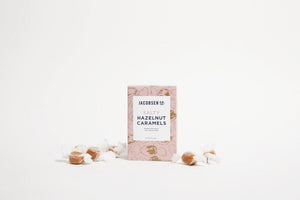 A rectangular pink box with brown floral patterns contains JACOBSEN SALT - SALTY HAZELNUT CARAMELS BOX by JACOBSEN SALT CO. The back of the box features descriptive text about the product's ingredients and origin, highlighting the use of Oregon sea salt and Jefferson hazelnuts for a rich flavor.