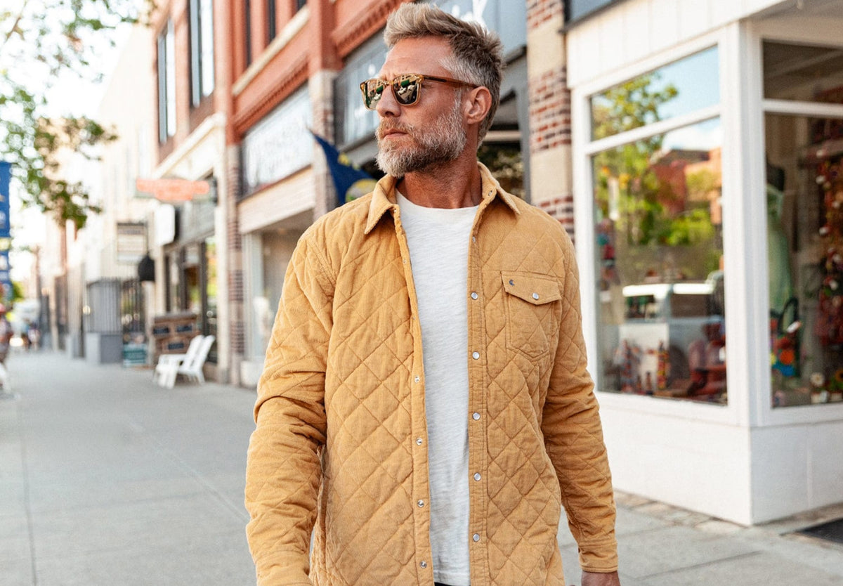 A man with short, curly hair and a beard wears the CRIQUET - CORDUROY QUILTED SHACKET by CRIQUET and blue jeans, standing against a white background while smiling and looking to his right.