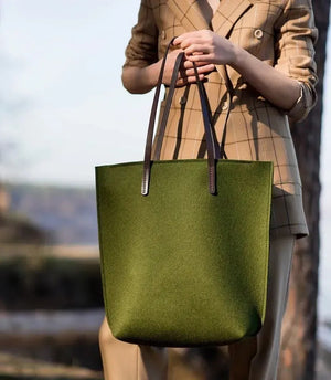 Close-up of hands in a beige knit sweater holding the WOOLBERRY WOOL FELT LONG TOTE BAG, featuring black leather straps and silver hardware.
