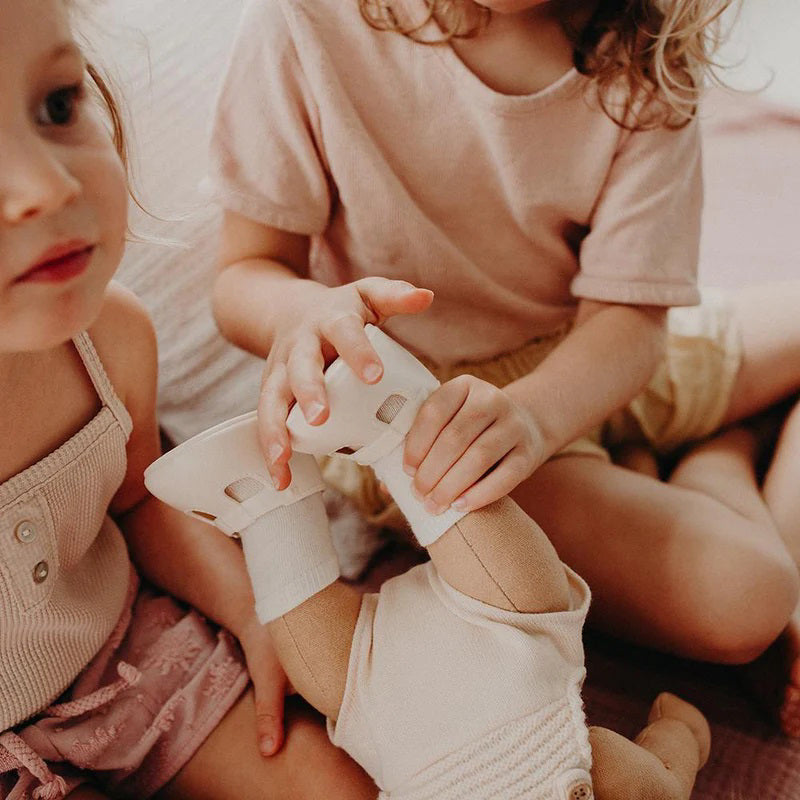A close-up of a baby wearing OLLI ELLA USA DINKUM DOLL SHOES in Mallow Pink and white socks. The baby is seated on a soft, pale pink textured blanket, dressed in light brown clothing. Only the lower half of the baby&#39;s body is visible.