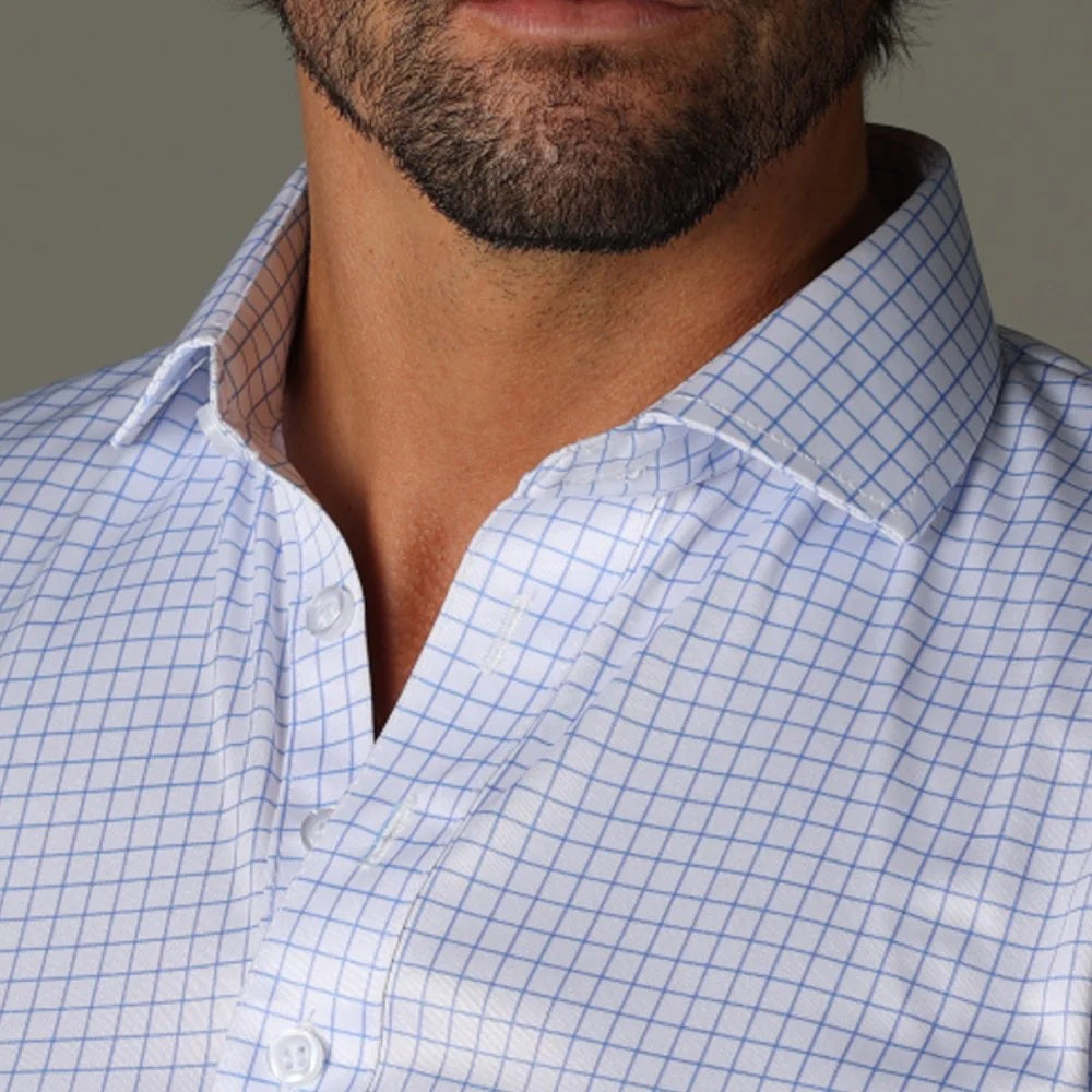 A close-up of a man wearing the COLLARS AND CO Semi Spread Collar Polo in Think Blue Check, featuring a comfortable white and light blue grid pattern with an open collar that reveals part of his beard and neck.