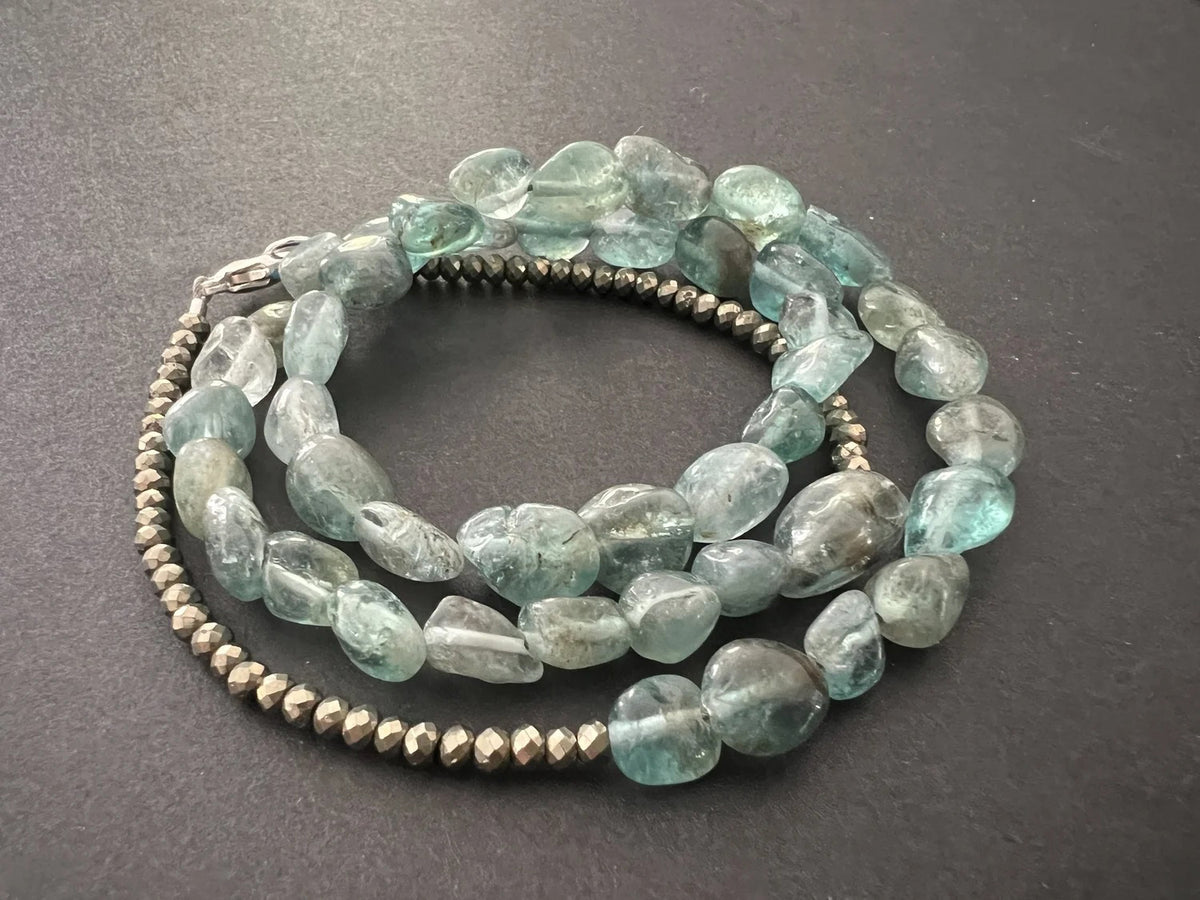 The ANN LIGHTFOOT - APATITE NUGGET TRIPLE WRAP BRACELET by ANN LIGHTFOOT features irregular light blue-green apatite stones, metallic round beads, and a sterling silver clasp, coiled in a circle on a dark surface.