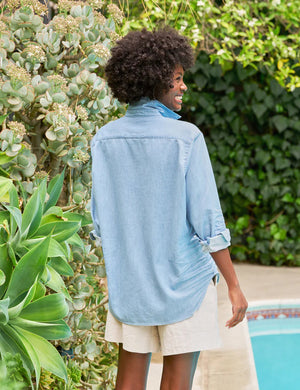 The FRANK & EILEEN Eileen Relaxed Button Up in Famous Denim Classic Wash is a light blue, long-sleeve shirt with bust-flattering buttons, a chest pocket, rolled sleeves, and a curved hem, shown against a white background.