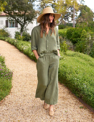 A woman wearing an olive green Tuscany Getaway Set, featuring the FRANK & EILEEN Maisie Wide Leg Linen Pull-On Pant, and a wide-brimmed straw hat strolls along a gravel path in a lush garden. With her hands in her pockets and a relaxed expression, she looks to the side, enjoying the serene ambiance.