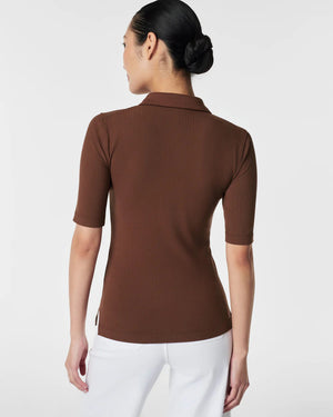 A woman in the SPANX - STRETCH RIB POLO TOP, a brown, ribbed, short-sleeve shirt with a button-down front, stands with her arms relaxed. The plain white background accentuates the top's stretchy ribbed fabric and body-hugging fit.