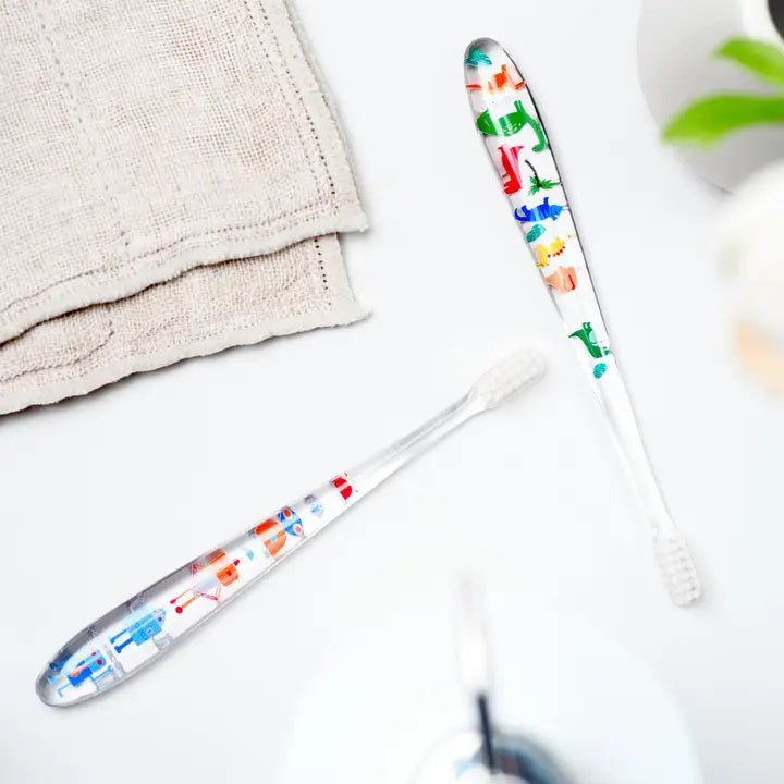 Two HAMICO - KIDS DINO TOOTHBRUSHES by HAMICO/INNOVATIVE TOOTHBRUSH, featuring unique dinosaur designs, sit on a white countertop next to a beige towel. A hint of a green plant is visible in the upper right corner, adding a touch of natural decor to the scene.