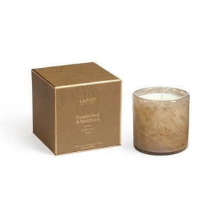 A candle in a transparent glass container, featuring a rich brown hue, is placed beside a coordinating brown box adorned with gold details. The set exudes a gourmand aroma and is elegantly labeled as "LAFCO - Pumpkin Seed & Sandalwood Candle 6.5oz." The candle's texture appears soft like fur.