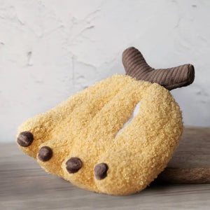 A small curly-haired gray dog is lying on a couch next to the LAMBWOLF COLLECTIVE - BANANA DOG TOY by LAMBWOLF COLLECTIVE, a soft, yellow plush toy shaped like a paw, complete with hidden squeakers. The dog appears relaxed and is looking directly at the camera.