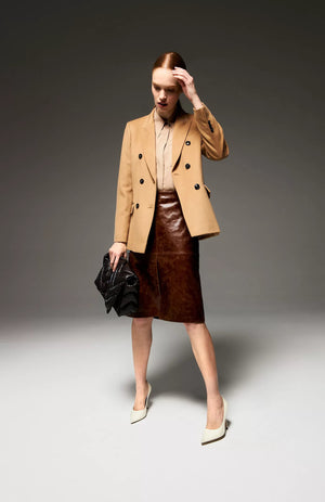 A person stands in a well-lit studio wearing a beige double-breasted blazer over a tan blouse, paired with the SEVENTY - VINTAGE EFFECT LEATHER PENCIL SKIRT by SEVENTY and white high-heeled shoes. They hold a black handbag made in Italy in one hand and have their other hand lightly touching their head.