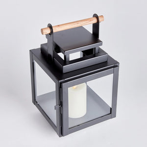 The ADWIN OUTDOOR LANTERN by NAPA HOME AND GARDEN is a black metal lantern with glass panels and a teak handle on top. Inside, there's a white pillar candle. The design is modern and exclusive, featuring clean lines and a sturdy structure, making it an ideal choice for both indoor and outdoor use.