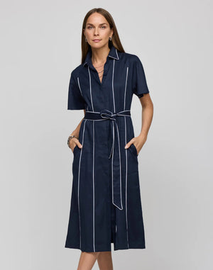 A woman with long brown hair wears the HINSON WU Joanna Dress, a navy knee-length piece featuring white piping and a waist tie belt made from stretch twill fabric, as she stands with hands in pockets against a plain background.