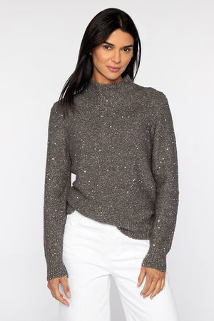 A person with long dark hair is wearing the KINROSS CASHMERE Sequin Funnel Neck Sweater, featuring a luxurious sparkly texture, paired with white pants. They are standing in front of a plain, light-colored background.