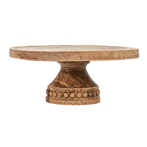 Introducing the Creative Coop "Mango Pedestal with Wood Beads," a wooden cake stand crafted from 100% mango wood. This elegant stand offers a round, flat surface perfect for holding cakes or desserts and features a decorative carved base adorned with circular bead patterns near the top. The natural finish of the mango wood beautifully showcases its unique grain and texture.