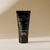 A black tube of PRESTON - HAND CREAM 3OZ with amber scent and shea butter, designed in a minimalist style, stands upright against a beige background. The 88 ml (3 oz.) tube features a hydrating formula and is cruelty-free, made in the USA. The product description is written in white text on the front.