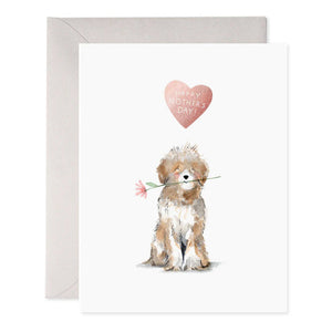 The DOG MOM MOTHERS DAY CARD by E.FRANCES PAPER features a handcrafted watercolor illustration of a brown dog sitting with its back turned, holding a pink flower in its mouth. This delightful image adorns a white greeting card, accompanied by a gray envelope in the background. Made in the USA, it beautifully encapsulates artistry and charm.