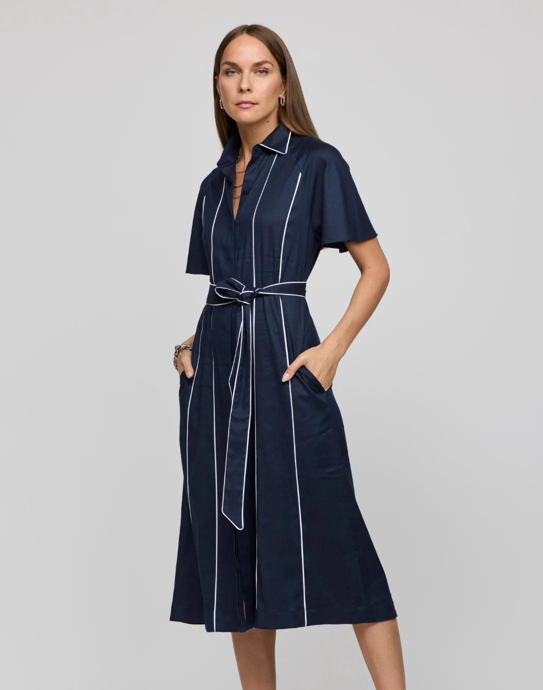 A woman with long brown hair wears the HINSON WU Joanna Dress, a navy knee-length piece featuring white piping and a waist tie belt made from stretch twill fabric, as she stands with hands in pockets against a plain background.