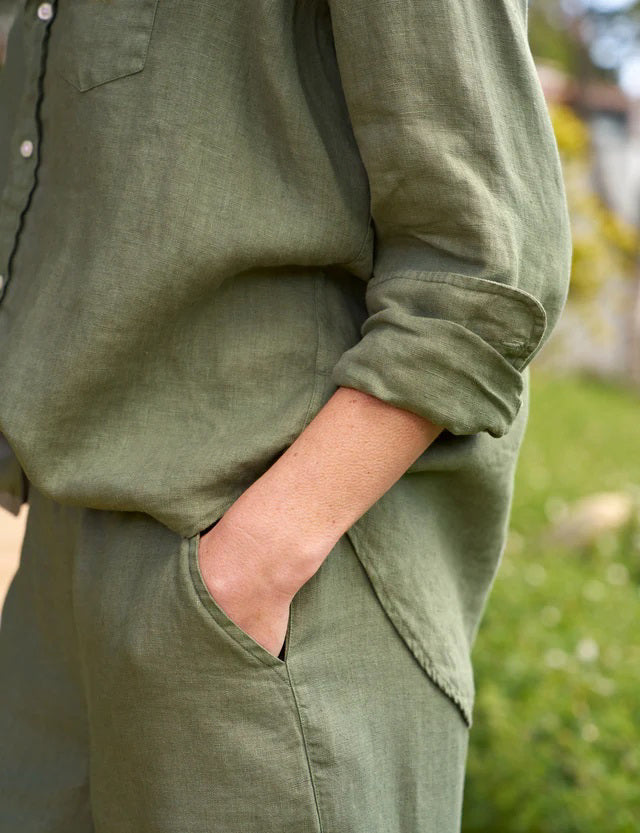 A person stands outdoors wearing an olive green FRANK AND EILEEN - EILEEN Relaxed Button Up Shirt in Linen from the Tuscany Getaway Set with the sleeves rolled to the forearms and matching Maisie pull-on pants. Their left hand is partially in their pocket, and greenery is visible in the background.