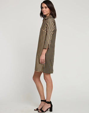 A woman stands posing sideways in a knee-length, loose-fitting HINSON WU AILEEN 3/4 Sleeve Dress featuring a classic black and tan stripe and gingham combo. She has shoulder-length dark hair and wears black high-heeled sandals. The background is a plain white wall.