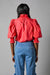 Rear view of a person wearing the Hope for Flowers Smocked Shirt, a bright red blouse with puffed sleeves, paired with blue jeans featuring pom-poms. Made from GOTS Certified organic cotton, this outfit embodies sustainable fashion against a gray background.