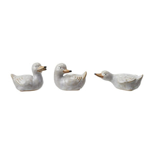 Three STONEWARE DUCKLING figurines by CREATIVE COOP sit on and in front of vintage books with worn edges, set against a plain white backdrop on a wooden surface, evoking nostalgia in their ceramic vintage reproduction form.