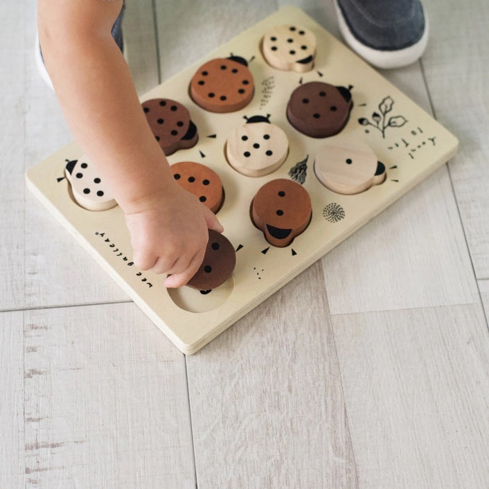 The WEE GALLERY - LADYBUGS COUNT TO 10 TRAY PUZZLE by WEE GALLERY is a wooden puzzle board featuring ladybug-shaped pieces in varying shades from light beige to dark brown. This educational puzzle helps teach counting skills and depicts leaves and twigs alongside the words &quot;Count to Ten&quot; on the left side. Each ladybug is adorned with black dots on their backs to represent number concepts.