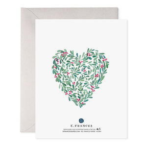 Introducing the HOLLY HEART HOLIDAY CARD by E.FRANCES PAPER: This exquisite greeting card showcases a heart-shaped design of green leaves and red berries, with "LOVE" beautifully centered. Crafted on luxurious paper, it is accompanied by a light gray envelope that evokes the charm of a delicate watercolor painting.