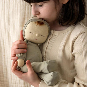 An adorable DOZY DINKUM DOLL by OLLI ELLA USA, featuring soft brown fabric skin, closed embroidered eyes, and short black hair, is dressed in a charming yellow onesie adorned with a rainbow design on the chest. Perfect as a first toy, the doll is lying on a white, quilted surface.