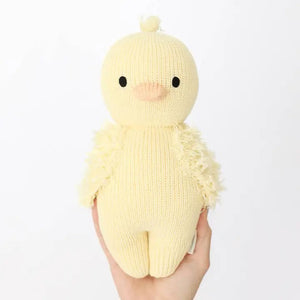 A soft, knitted yellow plush toy named "CUDDLE + KIND - HANDMADE BABY DUCKLING" by the brand CUDDLE & KIND, featuring a round head and body and viewed from the back. This charming toy has small wings and a tuft of 100% cotton yarn on its head, resembling a hand-knit baby chick while providing fair trade income to artisans.