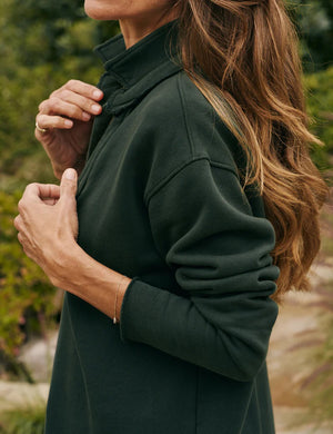 A person with long brown hair wearing a dark green FRANK & EILEEN - Patrick Popover Henley in Triple Fleece Evergreen stands outdoors. They are adjusting the collar of their top with both hands. The background is lush with greenery and soft-focus plants.
