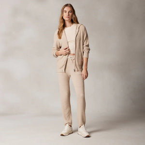 A person stands against a neutral background wearing a beige, coordinated outfit made from Kash-ease yarn. The ensemble consists of the KASHWERE - RELAXED JOGGER sweatpants, a zip-up hoodie, and a crop top. Paired with white sneakers, the easy-care durability of the ensemble is evident as they hold the hoodie with one hand.