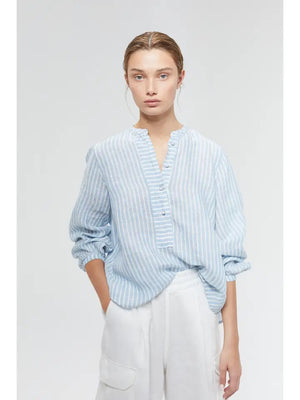 A person with light brown hair pulled back is wearing an oversized LANHTROPY - SAKE LINEN SHIRT by LANHTROPY, made of organic linen fabric, featuring a light blue and white striped pattern with buttons down the front and a mandarin collar. The sleeves are rolled up, and they have their left hand in the pocket of their white pants. The background is plain white.