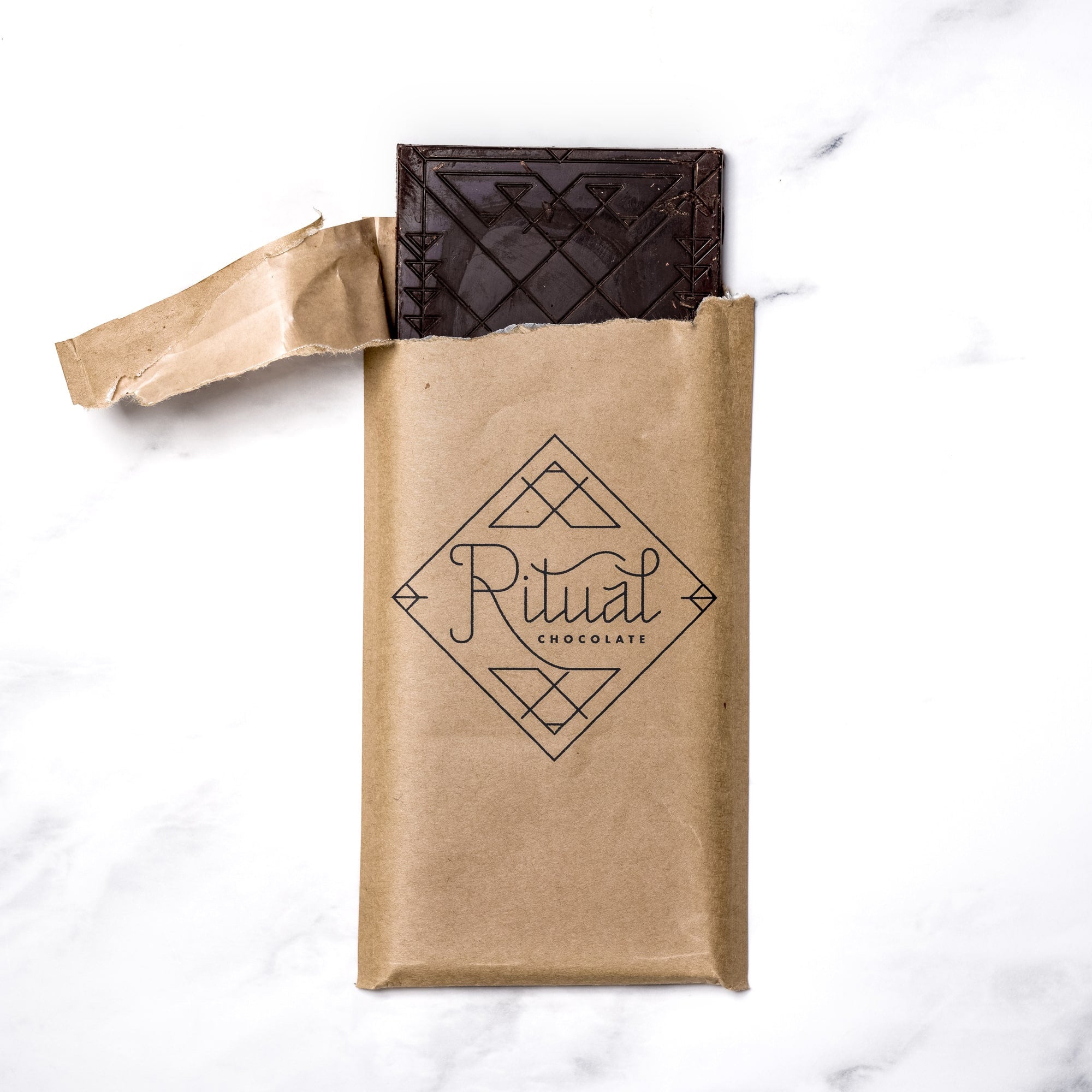 An opened brown paper package partially reveals a dark chocolate bar with intricate patterns. The package features a geometric design and the word "Ritual" written in a stylized font, followed by "One Hundred Percent Cacao Blend Chocolate Bar." The background is a white marble surface.