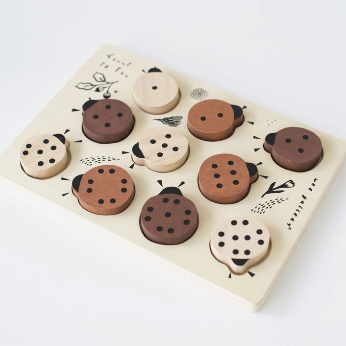 The WEE GALLERY - LADYBUGS COUNT TO 10 TRAY PUZZLE by WEE GALLERY is a wooden puzzle board featuring ladybug-shaped pieces in varying shades from light beige to dark brown. This educational puzzle helps teach counting skills and depicts leaves and twigs alongside the words &quot;Count to Ten&quot; on the left side. Each ladybug is adorned with black dots on their backs to represent number concepts.