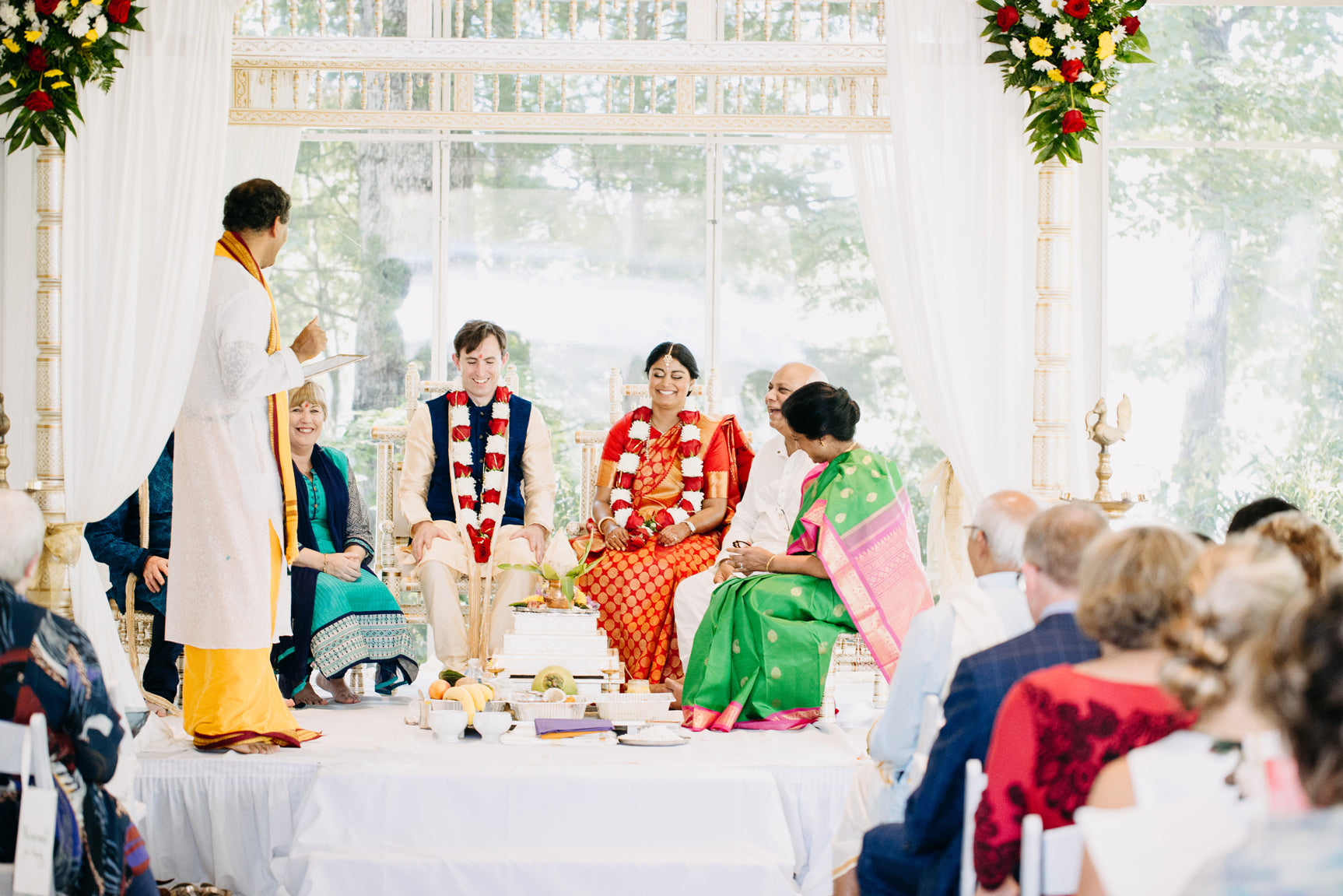 A Hindu wedding ceremony with the bride and groom seated at the altar, wearing traditional attire and flower garlands, surrounded by family and a priest, with guests watching. The setting is bright with floral decorations.