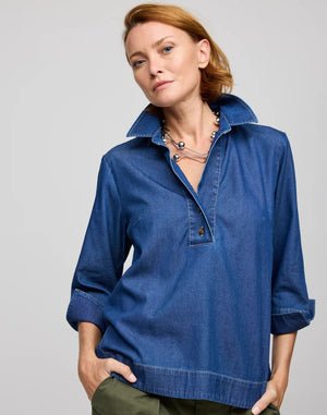 A person with short hair sits on a stool, facing away from the camera. They wear the breathable HINSON WU AILEEN 3/4 sleeve top in denim, featuring buttons down the back, paired with green pants.